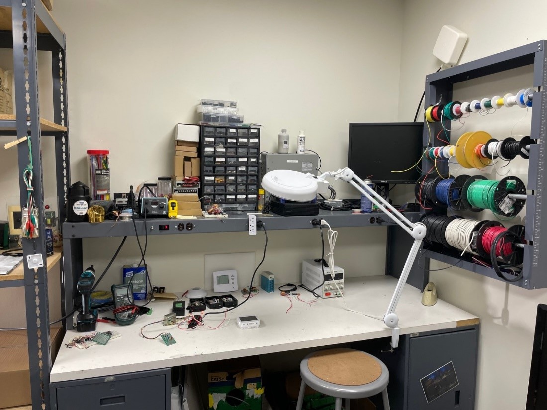 A cluttered electronics workshop with tools, spools of wires, monitors, and various electronic devices on a bench.