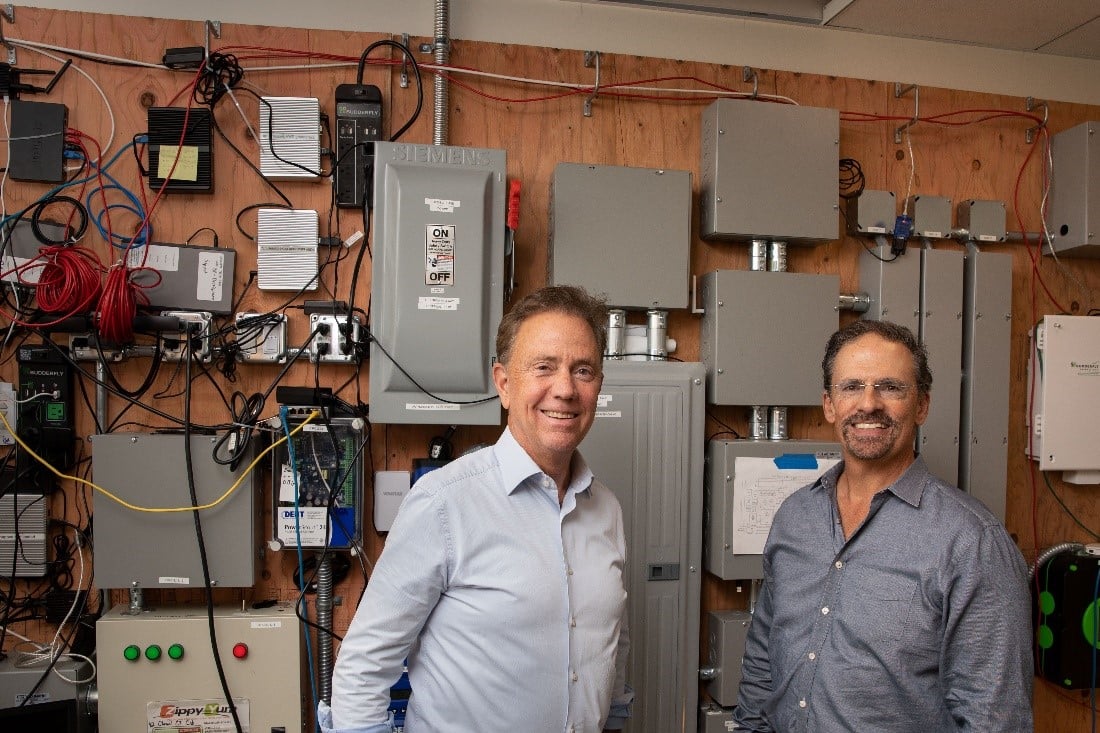 Two men smiling in a workshop with various electrical panels and equipment mounted on a wooden wall behind them.